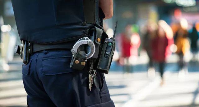 Police officer watching people. Detail Policeman with handcuffs and group of blurred people in background.